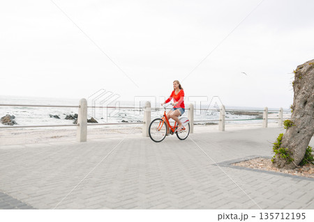 Female cyclist riding orange bicycle along seaside promenade by concrete railing with cloudy sky 135712195