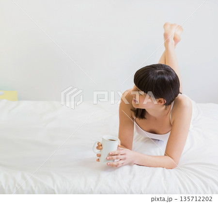 Woman lying on stomach holding white mug while wearing camisole on bed in bedroom, copy space 135712202