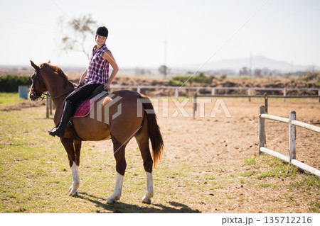 Female rider sitting astride chestnut horse in dirt paddock wearing helmet with burgundy saddle pad 135712216