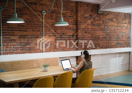 Adult woman wearing headset typing on laptop at table with succulent in office, copy space Adult woman wearing headset typing on laptop at table with succulent in office, copy space 135712234