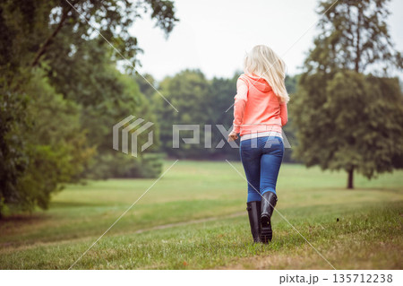 Woman running from camera across meadow wearing coral hoodie, blue jeans and black rubber boots Woman running from camera across meadow wearing coral hoodie, blue jeans and black rubber boots 135712238