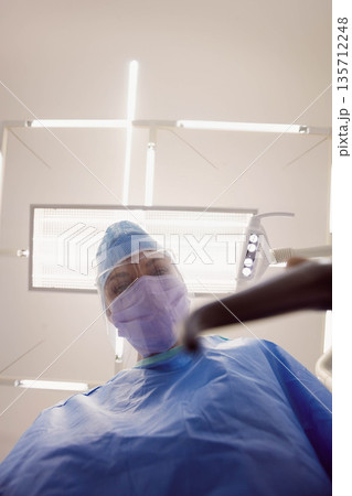 Female medical professional leaning beneath surgical light wearing mask, face shield in sterile OR 135712248