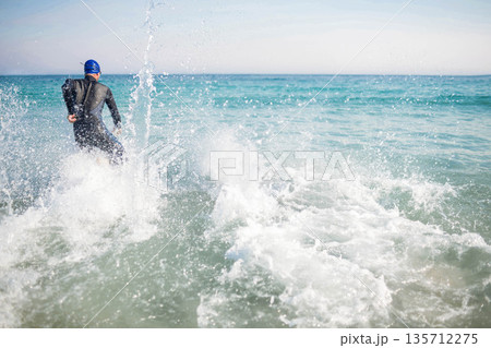 Male athlete striding into turquoise sea wearing neoprene wetsuit and blue swim cap, copy space Male athlete striding into turquoise sea wearing neoprene wetsuit and blue swim cap, copy space 135712275