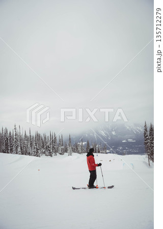 Male skier in red jacket standing on groomed slope by pines holding ski equipment, copy space Male skier in red jacket standing on groomed slope by pines holding ski equipment, copy space 135712279