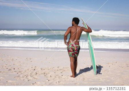 African American man standing shirtless on beach holding surfboard facing rolling ocean waves African American man standing shirtless on beach holding surfboard facing rolling ocean waves 135712281