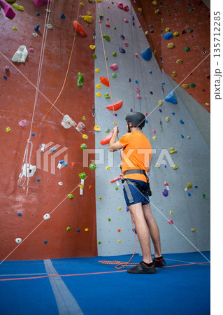 Adult male climber standing in gym wearing harness with ropes facing climbing wall, copy space Adult male climber standing in gym wearing harness with ropes facing climbing wall, copy space 135712285