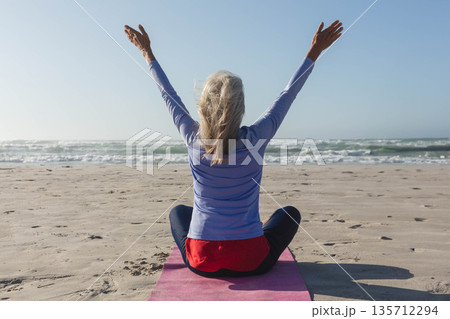 Senior woman practicing yoga sitting cross-legged on pink mat at oceanfront wearing athletic wear 135712294