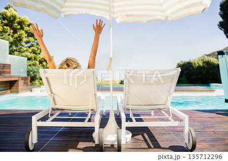 Adult female in swimsuit reclining under white beige striped umbrella by clear blue pool on deck Adult female in swimsuit reclining under white beige striped umbrella by clear blue pool on deck 135712296