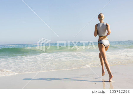 Woman walking into gentle waves on sandy shoreline wearing two-piece swimsuit, copy space 135712298