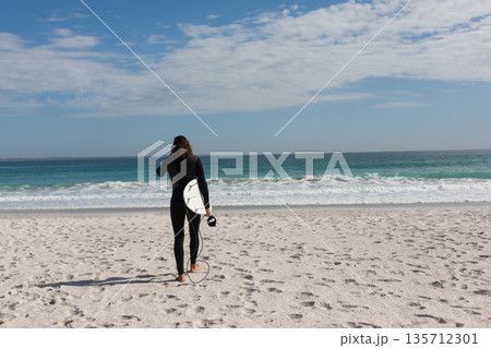 Female surfer in black wetsuit walking on beach carrying white surfboard and leash toward waves Female surfer in black wetsuit walking on beach carrying white surfboard and leash toward waves 135712301