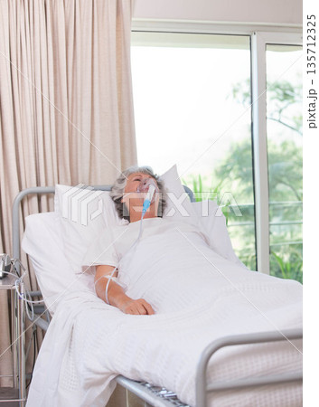 Senior female patient lying on hospital bed wearing hospital gown breathing with oxygen mask tubing 135712325