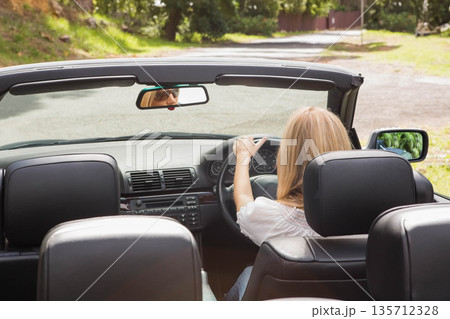 woman in mid-20s gripping steering wheel and driving convertible car on rural road under sunlight woman in mid-20s gripping steering wheel and driving convertible car on rural road under sunlight 135712328