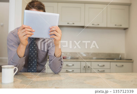 Adult male wearing tie holding tablet near face while sitting at kitchen countertop with coffee mug Adult male wearing tie holding tablet near face while sitting at kitchen countertop with coffee mug 135712339