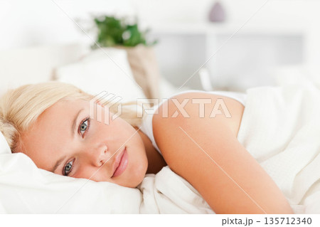Woman wearing white sleeveless top resting head on pillow and gazing at camera in calm bedroom Woman wearing white sleeveless top resting head on pillow and gazing at camera in calm bedroom 135712340