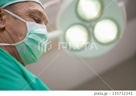 Senior male surgeon wearing green scrubs, mask, leaning under surgical light in OR copy space 135712341