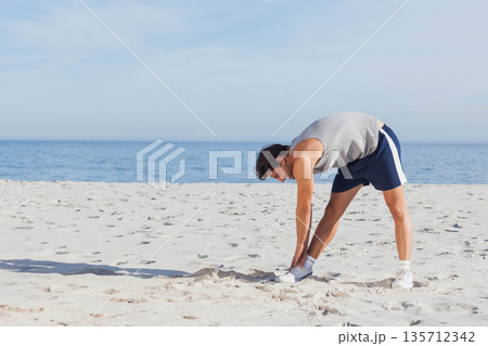 Man stretching forward in sleeveless top, sports shorts, athletic shoes on sandy beach, copy space Man stretching forward in sleeveless top, sports shorts, athletic shoes on sandy beach, copy space 135712342