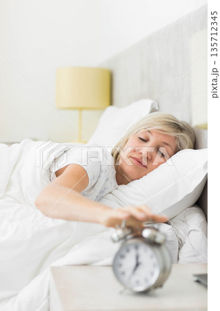 Mature adult woman wearing pajama top reaching analog alarm clock at bedside in bedroom Mature adult woman wearing pajama top reaching analog alarm clock at bedside in bedroom 135712345