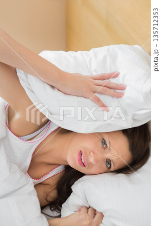 Woman holding pillow to ear lying in bed wearing pink tank top near wooden headboard 135712353