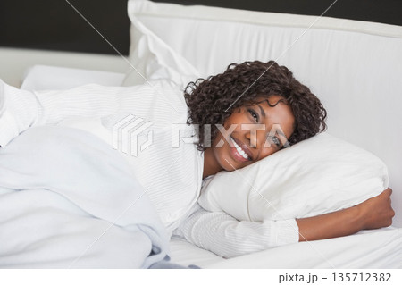 African American woman smiling and reclining on made bed in bedroom under blanket with pillow 135712382