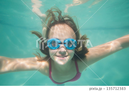 Female child swimming underwater in swimming pool wearing blue goggles and pink swimsuit top 135712383