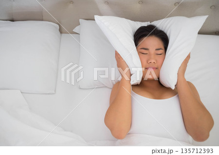 Asian woman reclining on bed pressing two white pillows against head with white bedding, headboard Asian woman reclining on bed pressing two white pillows against head with white bedding, headboard 135712393