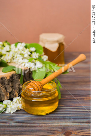 Sweet honey jar surrounded spring acacia blossoms. Honey flows from a spoon in a jar. jars of clear fresh acacia honey on wooden background 135712449