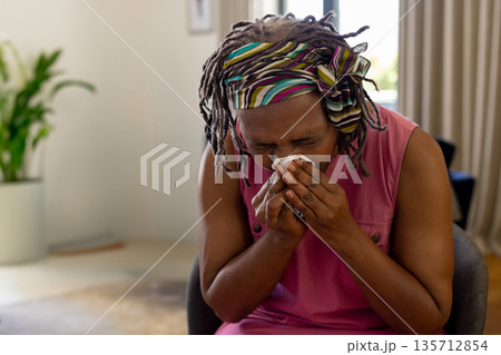 African American senior woman sneezing into white tissue while sitting at home near potted plant 135712854