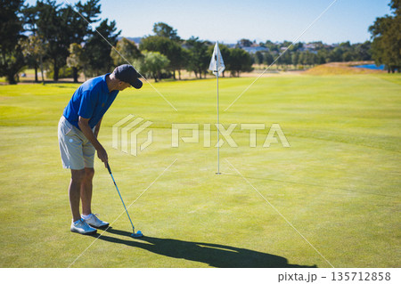 Senior golfer lining up putt on putting green with putter, golf ball near flagstick, copy space 135712858