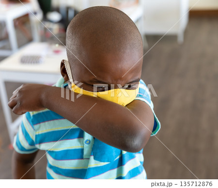 African American boy coughing into elbow, wearing yellow face mask in classroom with white tables African American boy coughing into elbow, wearing yellow face mask in classroom with white tables 135712876