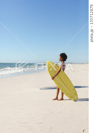 African American woman wearing bikini holding yellow surfboard facing waves on beach, copy space 135712879