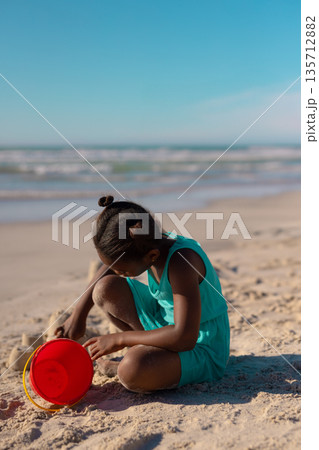 African American girl shaping sand structures on sandy shoreline using red bucket under clear sky 135712882