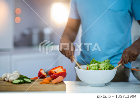 Mid adult African American man mixing salad in bowl in kitchen with wooden spoons, copy space 135712904