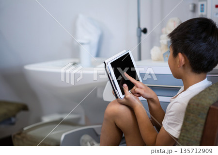 Male child sitting on padded chair and tapping blank tablet near porcelain sink and instrument cart Male child sitting on padded chair and tapping blank tablet near porcelain sink and instrument cart 135712909
