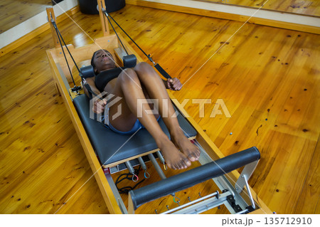 African American woman doing Pilates on wooden reformer using footbar with straps and springs in stu 135712910