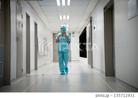 Female medical professional in scrubs walking down hospital hall with surgical cap and stethoscope 135712931