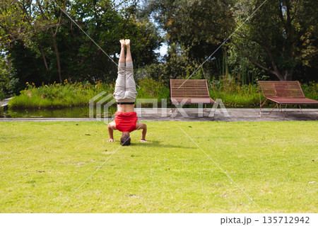 Adult male balancing headstand on lawn, wearing red tee beside wooden deck chairs overlooking pond 135712942
