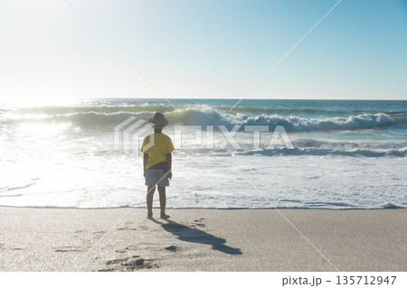 African American boy standing barefoot on beach wearing straw hat yellow T-shirt facing waves African American boy standing barefoot on beach wearing straw hat yellow T-shirt facing waves 135712947