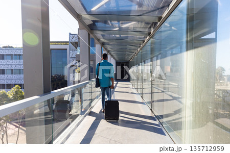 African American man walking through glass corridor pulling suitcase and carrying shoulder bag African American man walking through glass corridor pulling suitcase and carrying shoulder bag 135712959