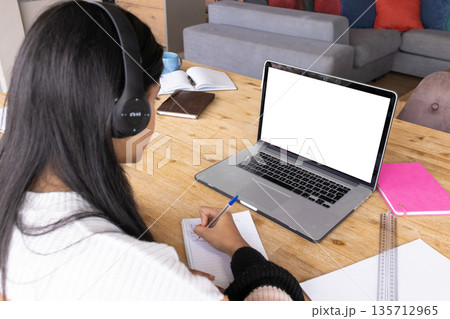 Teenage girl wearing white sweater black headphones writing in notebook at home study with laptop 135712965