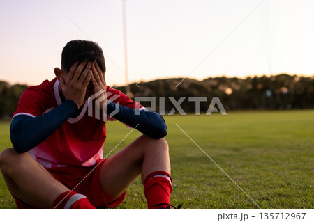 Teenage male soccer player sitting on grass field at dusk shielding face near floodlight poles 135712967
