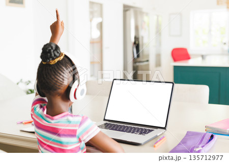 African American child student raising hand and sitting at kitchen table with laptop, headphones 135712977