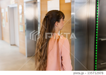 Mid-adult woman carrying tan leather bag standing in office lobby watching elevator green lights 135712978