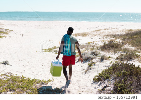 African American man walking across dune toward sea in trunks carrying green cooler, striped towel 135712991