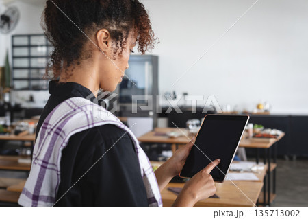 Mid adult woman tapping tablet while draping tea towel by wooden tables in restaurant, copy space Mid adult woman tapping tablet while draping tea towel by wooden tables in restaurant, copy space 135713002