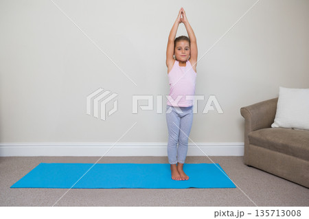 Female child performing full-body stretch on blue yoga mat at home with brown couch, white pillow 135713008