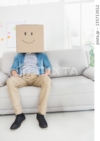 Sitting on light grey sofa in lounge, man wearing smiling cardboard box by flipchart, copy space 135713012