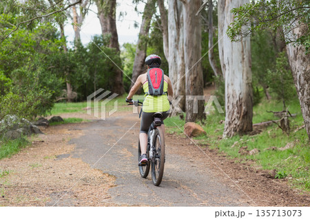 Female cyclist riding mountain bike along forest trail, wearing helmet and carrying hydration pack Female cyclist riding mountain bike along forest trail, wearing helmet and carrying hydration pack 135713073