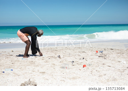 Senior African American man bending over on sandy beach holding trash bag picking bottles 135713084