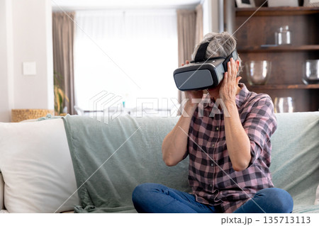 Senior woman wearing virtual reality headset and sitting on sofa in living room, copy space Senior woman wearing virtual reality headset and sitting on sofa in living room, copy space 135713113