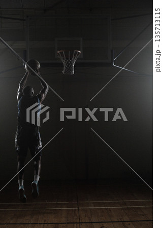 Basketball flying toward hoop in dimly lit gymnasium, spotlights casting dramatic shadows 135713115